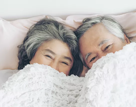 Senior asian couple laying in bed with the sheets covering half their faces.