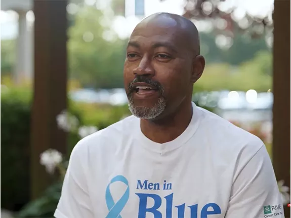 Terrance Gist, a prostate cancer patient in Williamsburg, VA, sitting down wearing a white shirt that has a blue prostate cancer ribbon that says "Men in Blue"