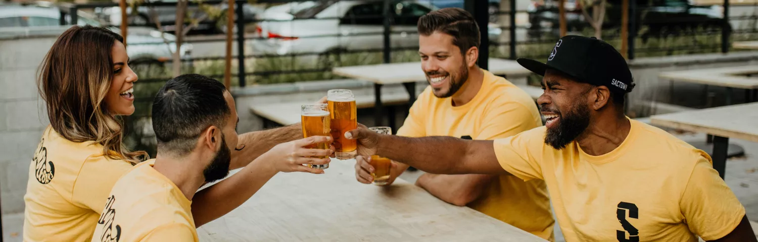 Four friends clink beers in yellow shirts at a table