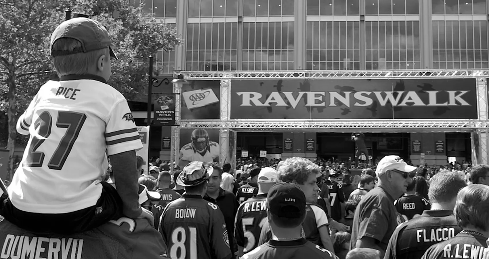 A black and white image of a crowd of people wearing football jerseys for the Baltimore Ravens, walking under an archway that says "RavensWalk." There is a focus on the left side of the image of a child sitting on an adult's shoulders.