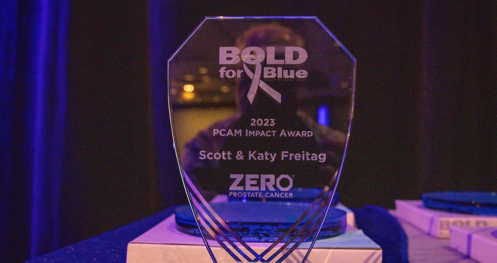 A glass award with the text "BOLD for Blue," "2023 PCAM Impact Award," "Scott & Katy Freitag," and "ZERO Prostate Cancer," set against a dark background.