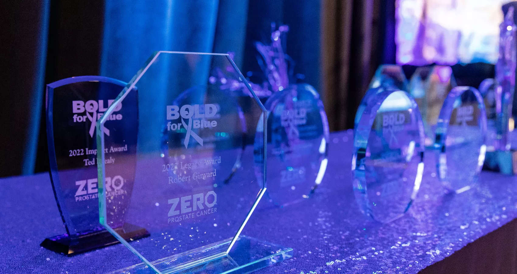 A collection of glass awards displayed on a sequined table. The awards are part of the "BOLD for Blue" initiative related to ZERO Prostate Cancer, including the 2022 Impact Award and Legacy Award. The setting appears to be a formal event.