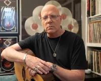 Jay Jay French of Twisted Sister with his guitar on his lap sitting in front of records hung on a wall