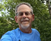 A senior man, Bob Jacobs, wearing a light blue t-shirt and taking a selfie in front of some greenery