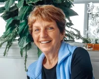 A woman with short reddish hair smiling while sitting in front of a window that has a large plant