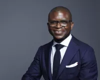 A black man wearing a blue suit against a gray background while sitting on a stool