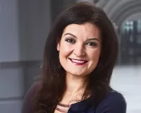 A person with long dark hair, wearing a dark top, is smiling and standing with arms crossed in a bright, modern hallway.