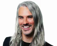 A person with long, silver hair and a beard, smiling broadly, wearing a black top against a plain white background.