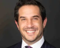 A person with short dark hair, wearing a dark suit, white shirt, and a patterned tie, smiling against a dark background.