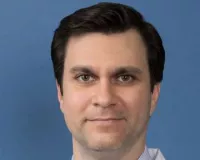 A person with short dark hair, wearing a white lab coat, a light blue striped shirt, and a patterned tie, smiling against a solid blue background.