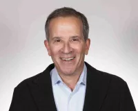 A person with short hair, wearing a black blazer over a light blue shirt, smiling against a light gray background.