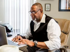 A man sits at a table with his laptop open, tuning into a virtual education session.