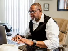 A man sits at a table with his laptop open, tuning into a virtual education session.