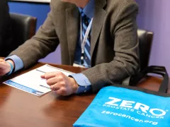 A person in a suit gestures while discussing materials on a table. A blue tote bag with "ZERO PROSTATE CANCER" is prominently displayed, highlighting prostate cancer awareness.