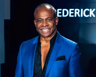 A middle aged Black man wearing a dark blue shirt with open buttons to his mid-chest and a royal blue jacket. 