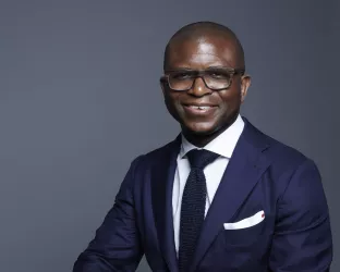 A black man wearing a blue suit against a gray background while sitting on a stool