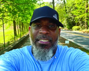 An older black man with a grey beard taking a selfie in a park wearing a ZERO Run/Walk shirt and hat