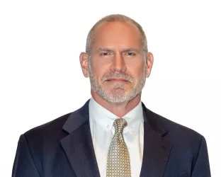 A bald, white man wearing a suit and tie, standing against a white backdrop
