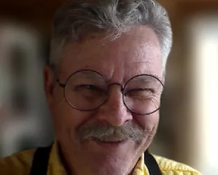 An older man with a gray mustache wearing round glasses, a yellow shirt, and black suspenders