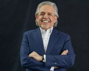 Luis Miranda Jr wearing a blue suit standing with his arms crossed against a black backdrop