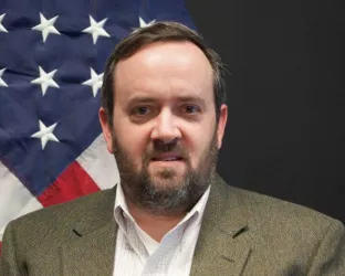 A man wearing a white shirt and green jacket sitting in front of an American flag