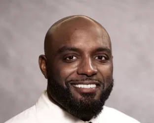 A bald Black man with a beard wearing a doctor's coat and tie for a headshot