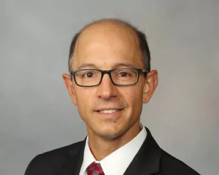A man with glasses, wearing a black suit with red tie, sitting for a headshot