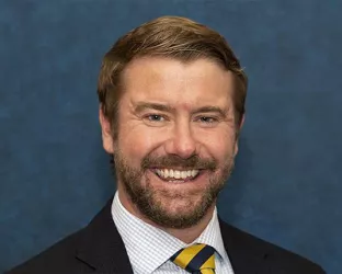 A middle aged white man with light brown hair and a beard smiling against a blue background