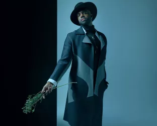 A man wearing a hat and trench coat holding flowers in an artsy way