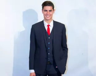 A young man with brown hair slicked to the side wearing a suit, best, and red tie smiling for cameras