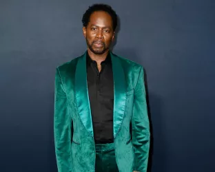 A Black man wearing a green suit and black shirt standing against a dark blue background