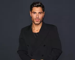 A man with olive skin, dark brown hair, and sharp features wearing a black peacoat standing against a dark blue background