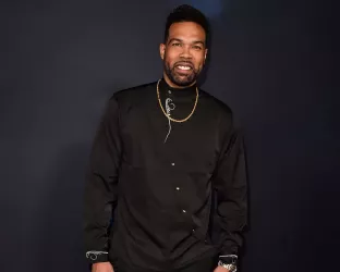 A Black man wearing a black shirt and gold necklace against a dark blue backdrop