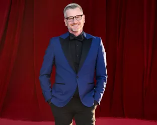 An older man wearing a blue suit and black pants standing against a red backdrop