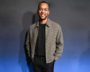 A young Black man wearing all black with a beige jacket