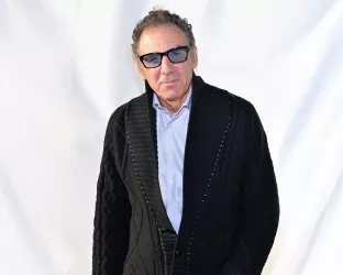 An older man with curly hair and glasses wearing a black cardigan