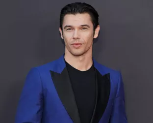 A young man with dark hair slicked back wearing a black tshirt with a blue and black suit jacket