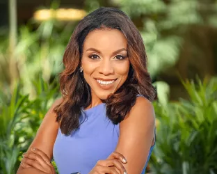 A Black woman in a purple top standing with her arms crossed and smiling in front of green foliage