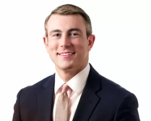 A person in a dark suit and light tie, smiling, with short hair, sitting against a plain white background.