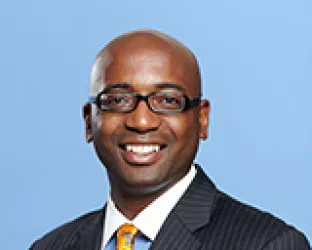 A person wearing glasses and a dark pinstripe suit with an orange and blue patterned tie, smiling against a light blue background.