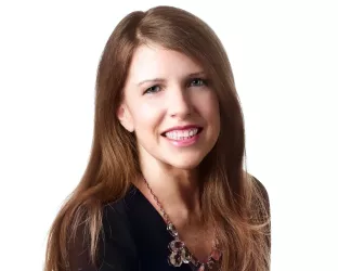 A person with long, light brown hair, wearing a black top and a necklace, smiling against a plain white background.