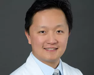 A professional headshot of a male doctor wearing a white lab coat embroidered with 'Urology Partners' and his name, along with a blue dress shirt and a plaid tie. He has short black hair and is smiling against a neutral gray background.
