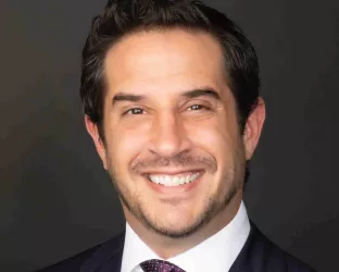 A person with short dark hair, wearing a dark suit, white shirt, and a patterned tie, smiling against a dark background.