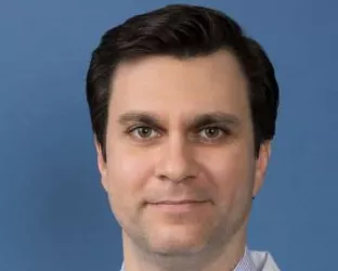 A person with short dark hair, wearing a white lab coat, a light blue striped shirt, and a patterned tie, smiling against a solid blue background.