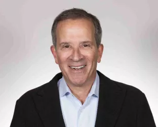 A person with short hair, wearing a black blazer over a light blue shirt, smiling against a light gray background.