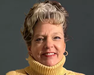 A professional portrait of a woman with short, wavy hair featuring blonde highlights, wearing a yellow turtleneck sweater and a heart-shaped pendant necklace. She is smiling warmly against a neutral gray background.