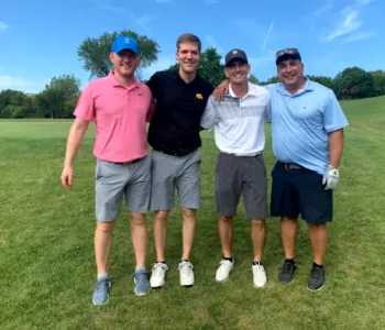 Four men in casual golf attire posing on a rolling golf green in front of trees