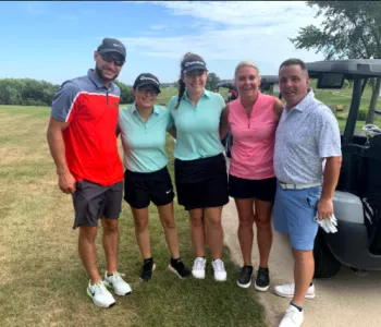 Three ladies stand inbetween two males all in golf attire on a golf course next to a luxury golf cart