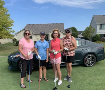 Four women in golf attire standing with drivers in front of a luxury gray vehicle on a golf course