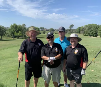Four older men sporting shading hats and different golf clubs stand on a rolling green in front of trees at a golf course